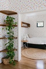 A room with white wallpaper adorned with colorful, scattered circles resembling icons often associated with social media platforms, creating a playful and lively atmosphere. The wallpaper complements the minimalistic, modern decor and is applied to both the walls of a bedroom and an adjacent hallway. This design brings a contemporary twist to traditional wallpaper.