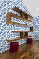 Wall covered with blue French palm leaf patterned wallpaper on a white background, with wooden shelves and red stools in a room with wooden flooring. Traditional wallpaper.