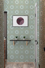 The wallpaper design has intricate mandala patterns in soothing shades of green, blue, and beige, with detailed geometric shapes and floral elements creating a harmonious and symmetrical look. The image also shows a small shelf with plants and a framed abstract art piece on the wall. Traditional wallpaper.