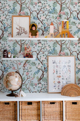 Self-adhesive wallpaper with a tree design, featuring intricate branches, delicate pink flowers, and green leaves set against a light blue background. The natural elements create a serene, nature-inspired look in the room, complementing the decor on the shelves and the wicker baskets below.