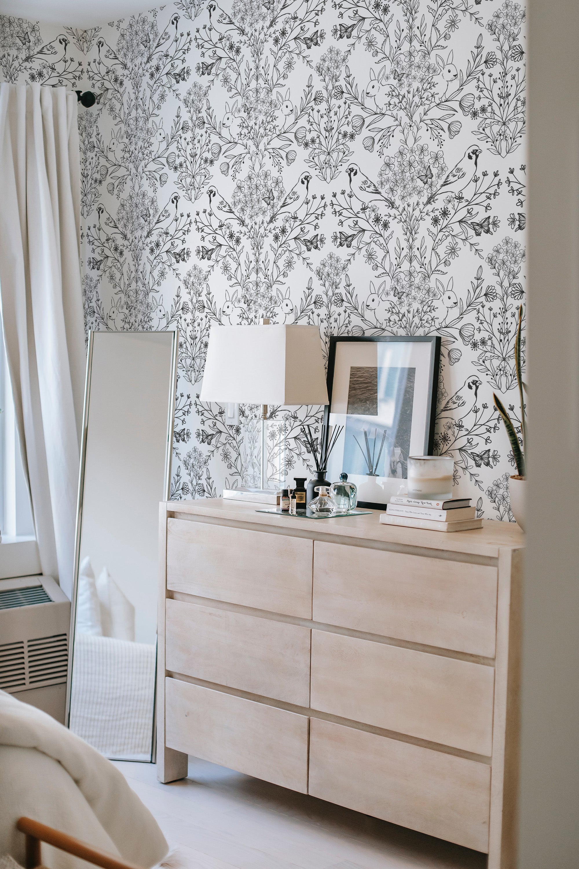A room with a black and white wallpaper design intricately intertwined with birds and floral patterns covering the wall behind a wooden dresser, evoking a classic and traditional wallpaper.