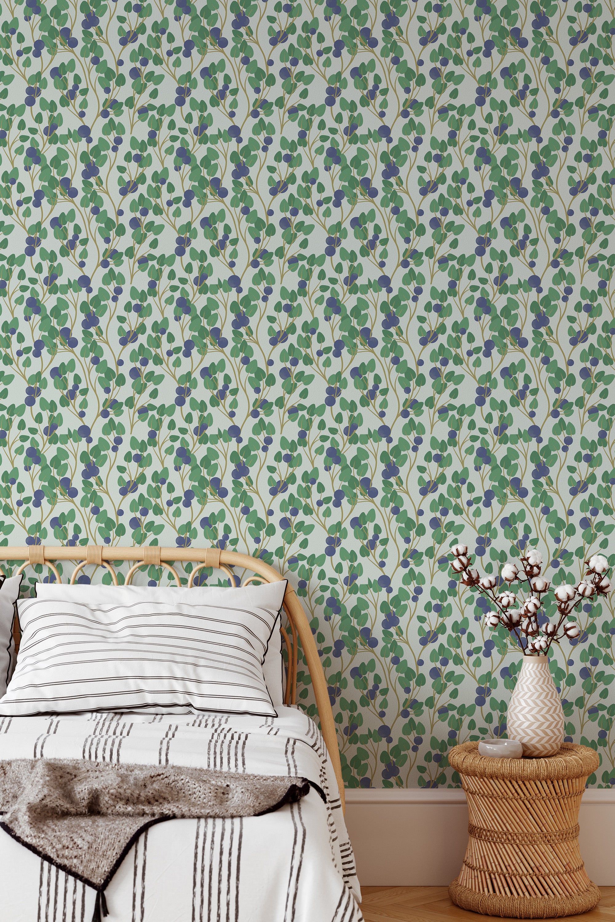 Self-adhesive wallpaper in a vibrant blueberry pattern with clusters of blue berries and green leaves on a white background, adorning the wall behind a cozy bed with striped bedding and a wicker side table.
