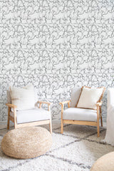 A room with a wall covered in wallpaper characterized by a swirling abstract marble texture in soft hues of white, gray, and hints of pink, providing a harmonious and calming aesthetic. Two light wood armchairs with white cushions are placed in front of the wall, with a woven ottoman and a plush patterned rug on the floor. Traditional wallpaper.