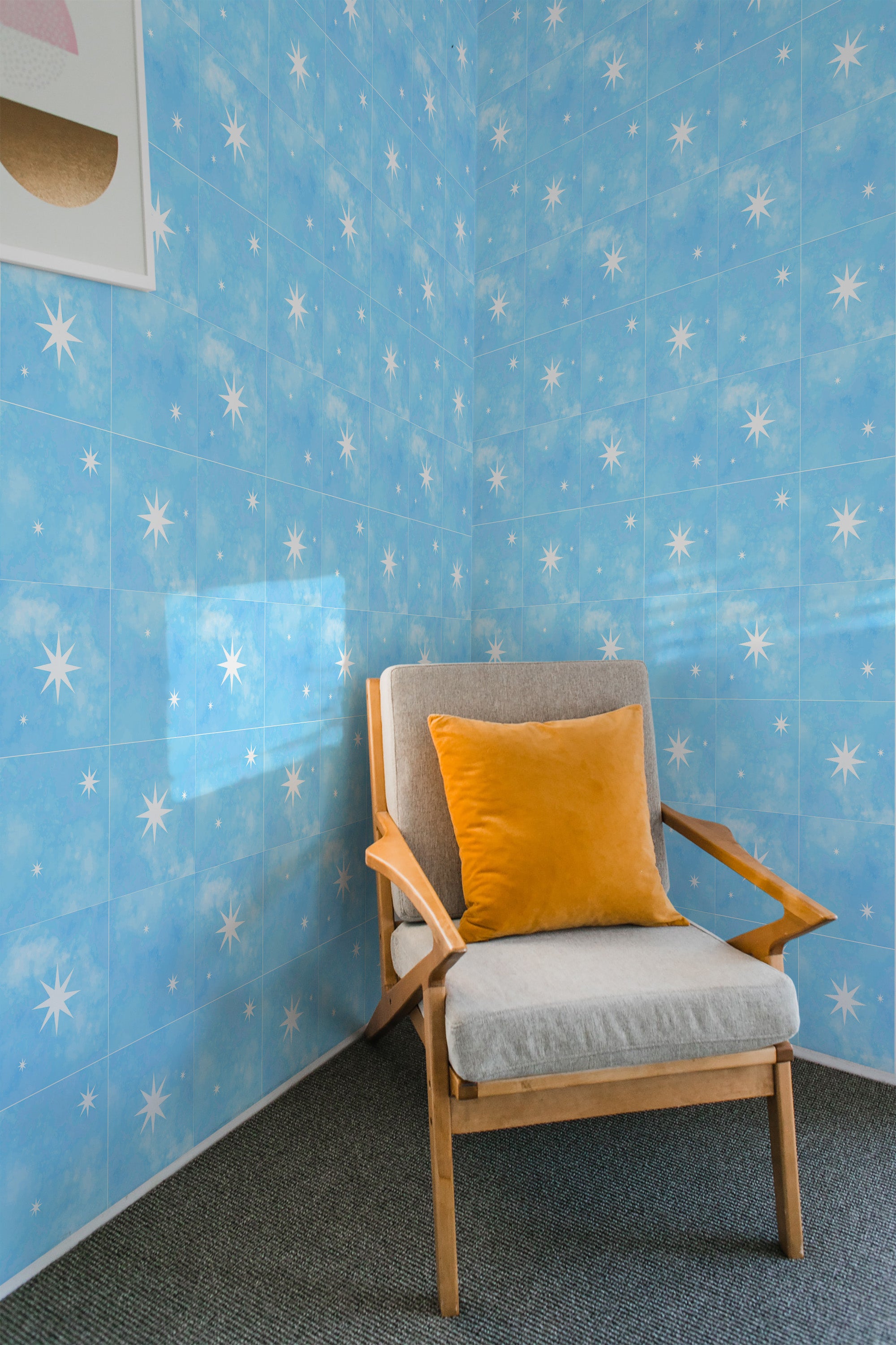 A room with light blue wallpaper adorned with white star-shaped patterns arranged in a tile-like grid. The wallpaper has a calming, sky-like appearance with the stars adding a touch of whimsy, creating a serene atmosphere. A wooden chair with a gray cushion and mustard-yellow pillow is placed against the wall. Traditional wallpaper.
