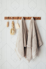 Self-adhesive minimalist wallpaper with a geometric line art pattern in light, neutral tones, rectangular shapes intersect throughout, creating a subtle, modern visual interest on the wall behind a wooden towel rack with hanging beige towels and rustic bath accessories.