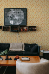 Yellow tulips pattern covers the wallpaper with small orange flowers scattered evenly, creating a lively and warm ambiance. The design blends well with the cozy and inviting decor of the room, complementing the black couch and wooden coffee table. Traditional wallpaper.