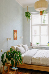 A cozy bedroom with walls adorned in a light wash denim wallpaper, creating a subtle, textured look with a soft blue and gray pattern. The design gives the room a calm and relaxed ambiance, complemented by natural light coming from a large window and hanging plants that add a touch of greenery. The wallpaper adds elegance to the space, making it a perfect blend of modern and traditional wallpaper.