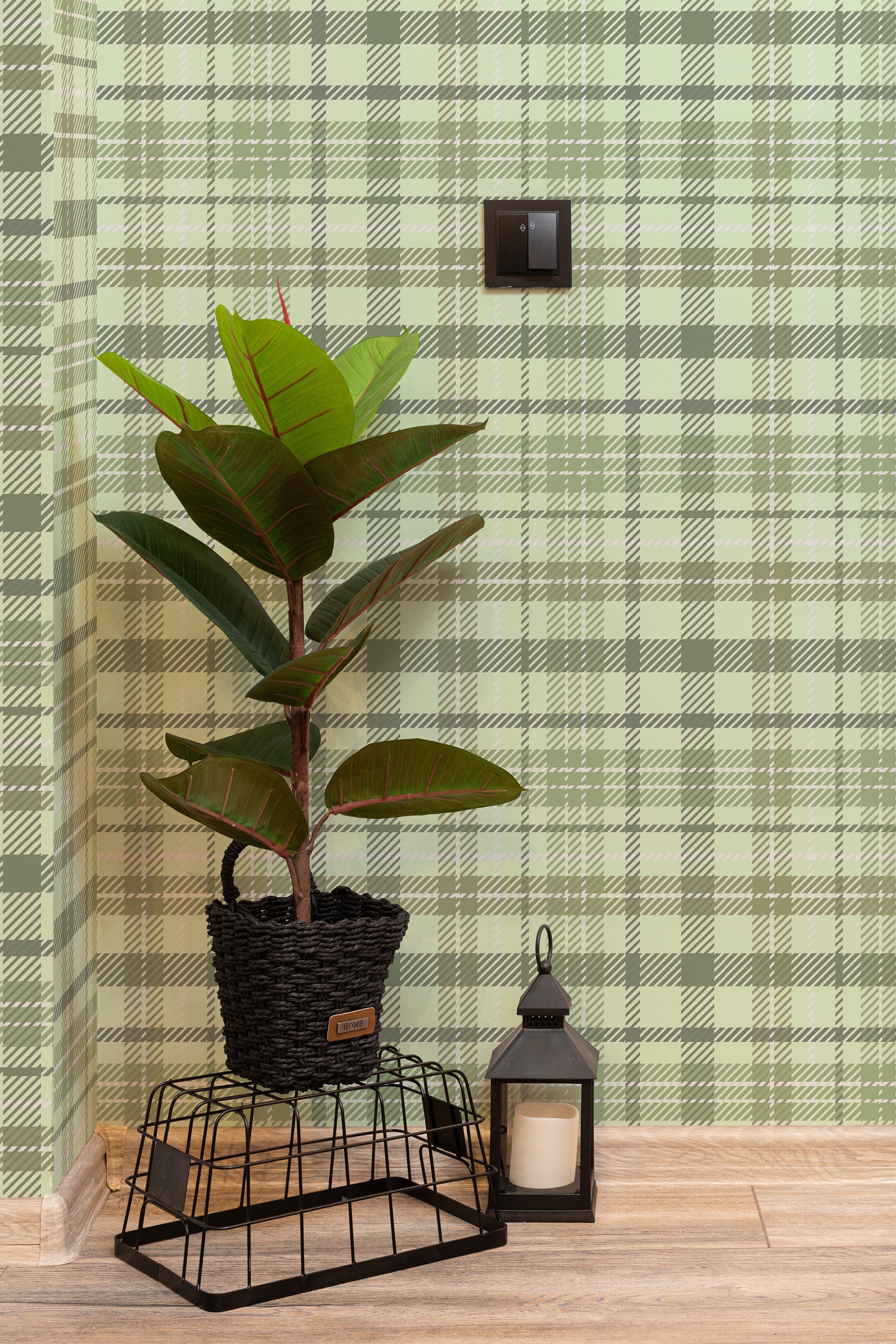 Self-adhesive wallpaper with a green plaid pattern and light, crisscrossing lines, giving a textured, woven appearance. The wall also has a black light switch, and a potted plant with broad leaves sits on a metal stand beside a decorative lantern on a light wood floor.