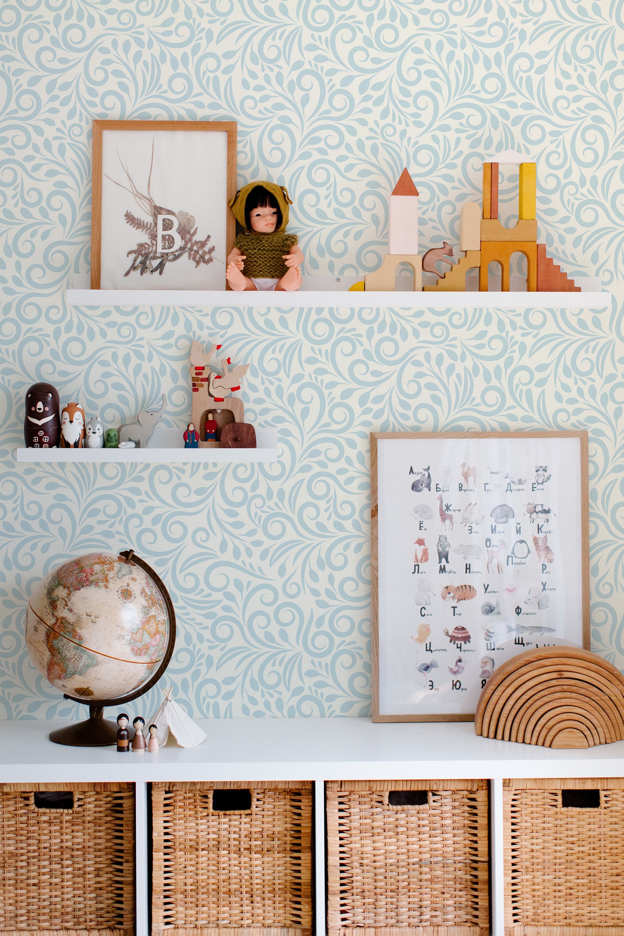 Self-adhesive wallpaper with a light blue intricate ornamental pattern, featuring flowing swirls and curls, covering the entire wall behind a shelf filled with decorative items, including a globe, toys, and framed artwork.