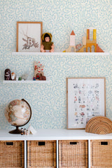 Self-adhesive wallpaper with a light blue intricate ornamental pattern, featuring flowing swirls and curls, covering the entire wall behind a shelf filled with decorative items, including a globe, toys, and framed artwork.