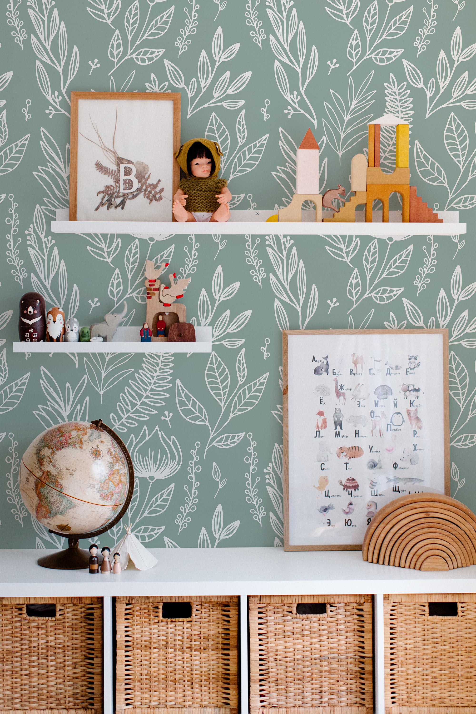 Self-adhesive sage green nursery wallpaper with a pattern of white leaves and branches, complemented by wooden shelves with toys and framed pictures.