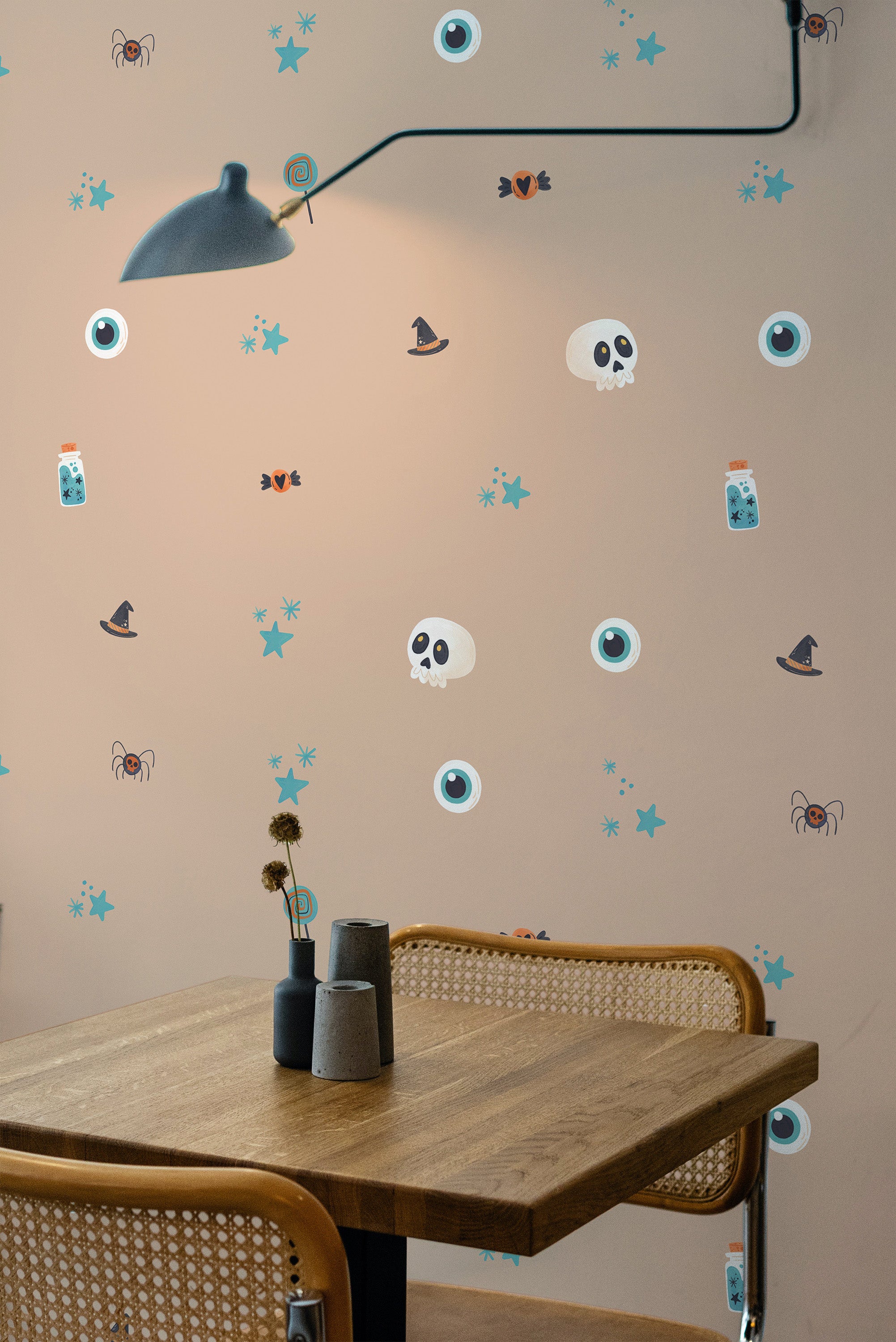 Self-adhesive Halloween wallpaper with skulls, witch hats, bats, and eyeballs on a neutral background in a room with a wood table and lamp.