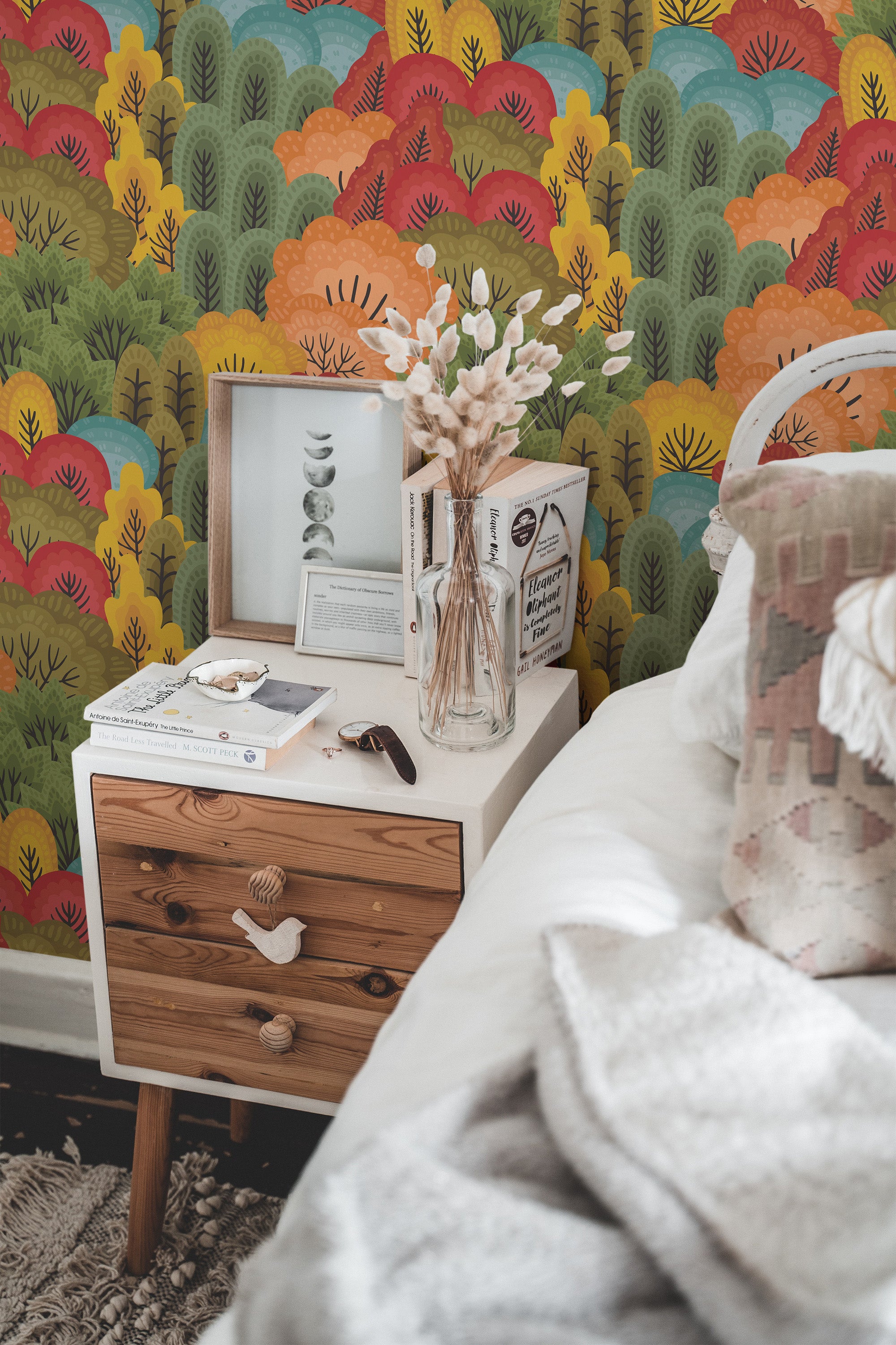 Self-adhesive wallpaper with a colorful pattern resembling autumn foliage in a forest, including red, orange, yellow, and green leaves, applied to a room wall behind a wooden bedside table and bed.