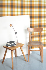 Self-adhesive Orange autumn plaid wallpaper pattern with warm shades of brown and beige, installed on a room's wall behind a wooden chair and table setup.