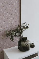 Self-adhesive wallpaper with a pattern of pinkish delicate flowers and subtle green leaves on a beige background, partially covered by a white canvas and accompanied by green vases with foliage.