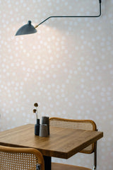 Self-adhesive Funky Dots wallpaper with a pattern of staggered white circles on a taupe background, applied in a room with a wooden table and wall-mounted lamp.