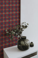 Self-adhesive burgundy plaid wallpaper on a wall with a white shelf holding green vases and foliage.