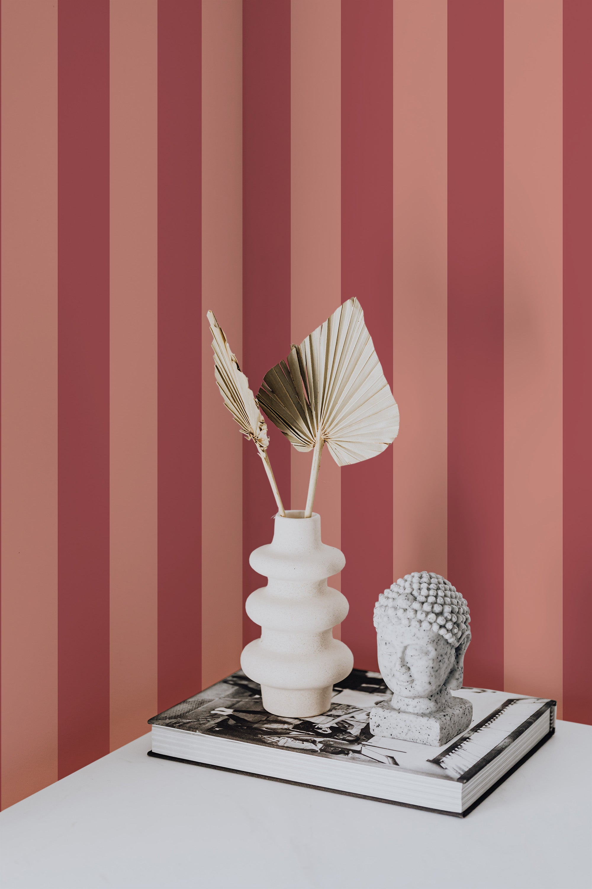 Self-adhesive wallpaper with alternating pink and red vertical stripes on a room wall, behind a white sculptural vase with leaves and a decorative object on top of a book.