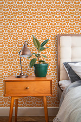 Self-adhesive wallpaper with an orange and white geometric scandi leaf pattern on a bedroom wall, complementing a wooden nightstand, a green potted plant, and a copper lamp.