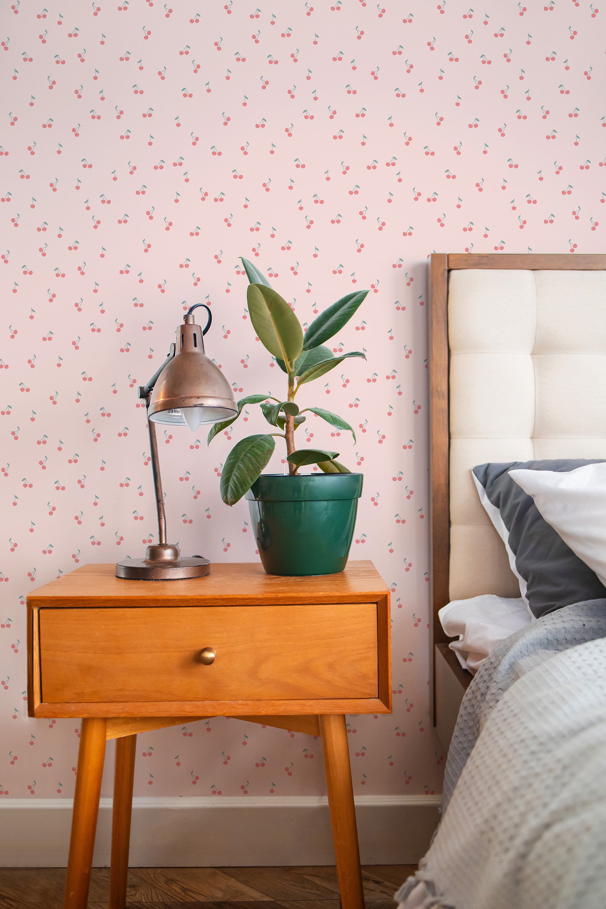 Self-adhesive wallpaper with a girly cherry pattern on a pink background, complementing a cozy bedroom interior with a wooden bedside table, lamp, plant, and bed.