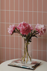 Self-adhesive pink tile-patterned wallpaper with lines creating a grid design on a wall, complemented by a bouquet of pink flowers on a table.