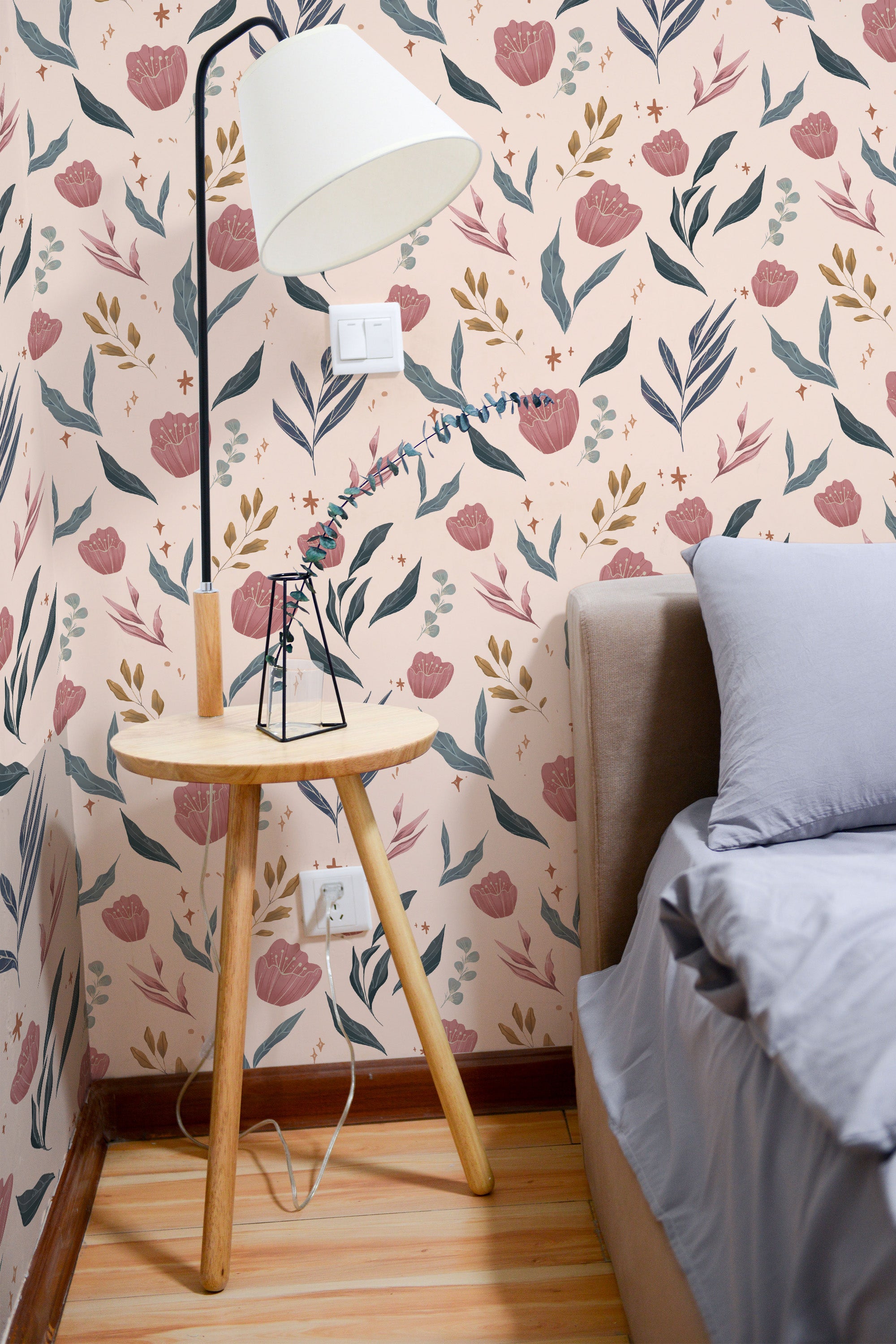 Self-adhesive Peachy Bouquet wallpaper with a pattern of pink and blue flowers and green foliage on a cream background in a room with a bed and a wooden side table.