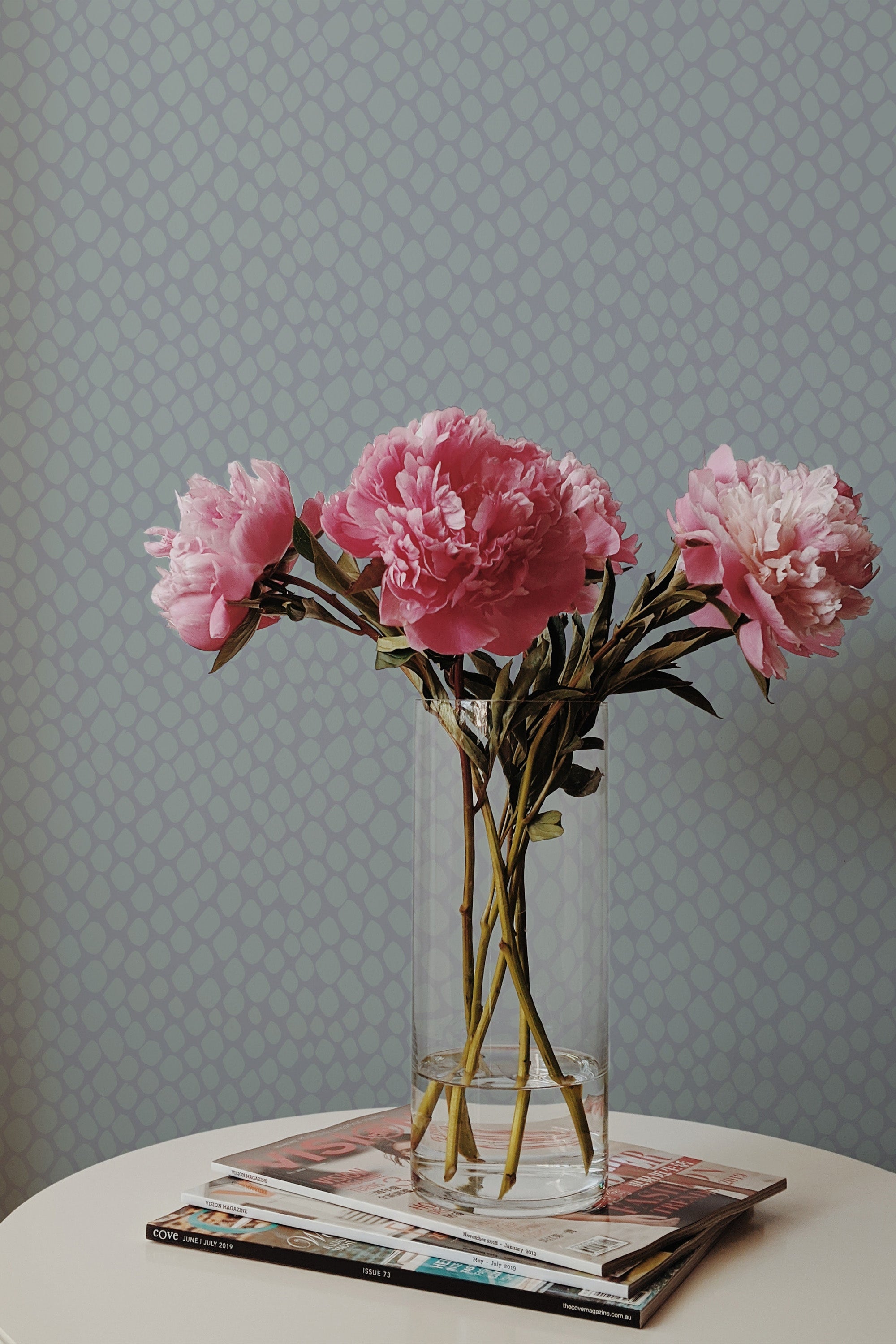 Self-adhesive wallpaper with a light blue background and a small geometric pattern behind a vase of pink peonies on a table.