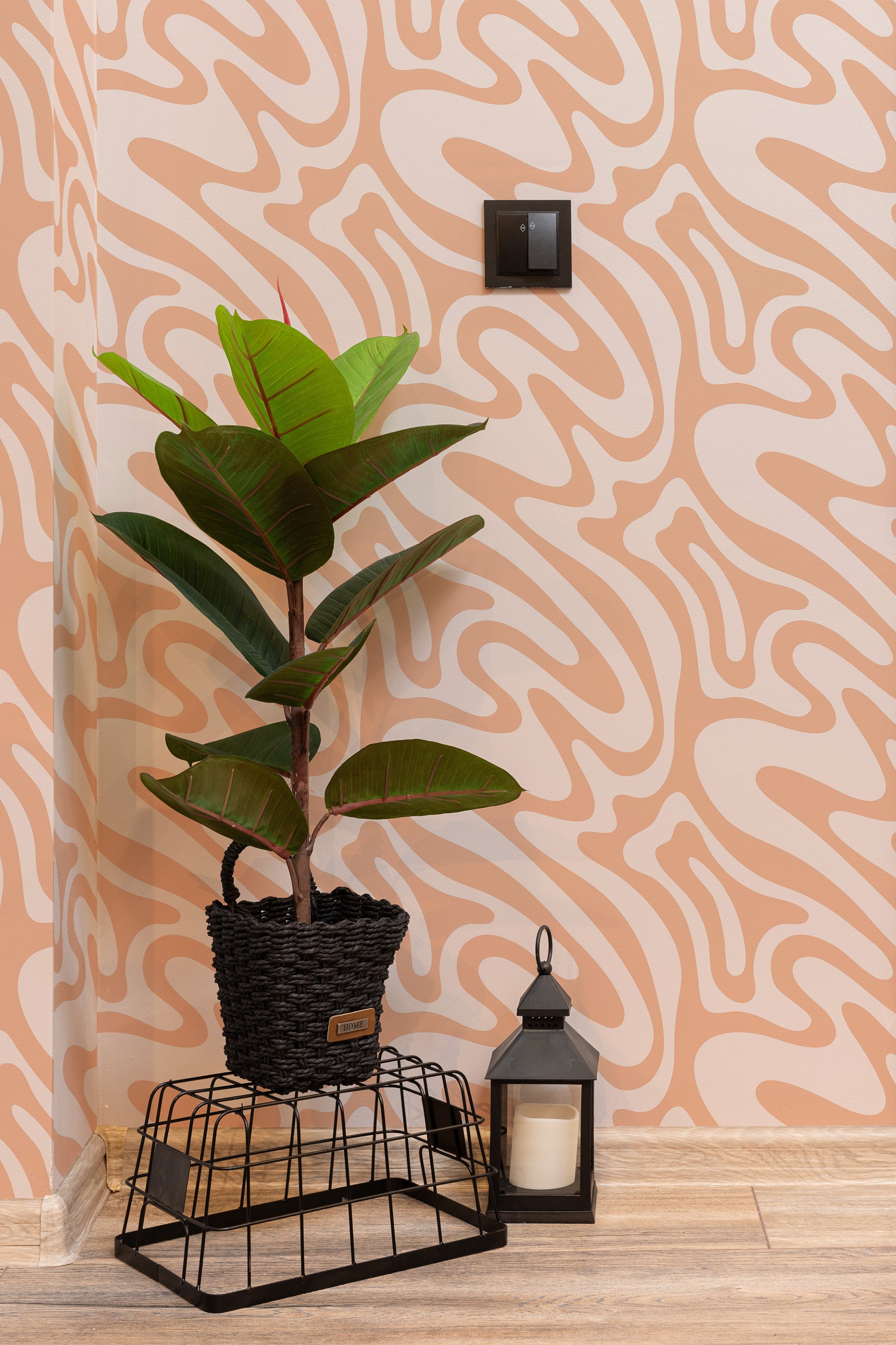 Self-adhesive Peach Funky Wave wallpaper with abstract wavy lines in shades of peach and cream, accompanied by a potted plant, a lantern, and a wall switch.