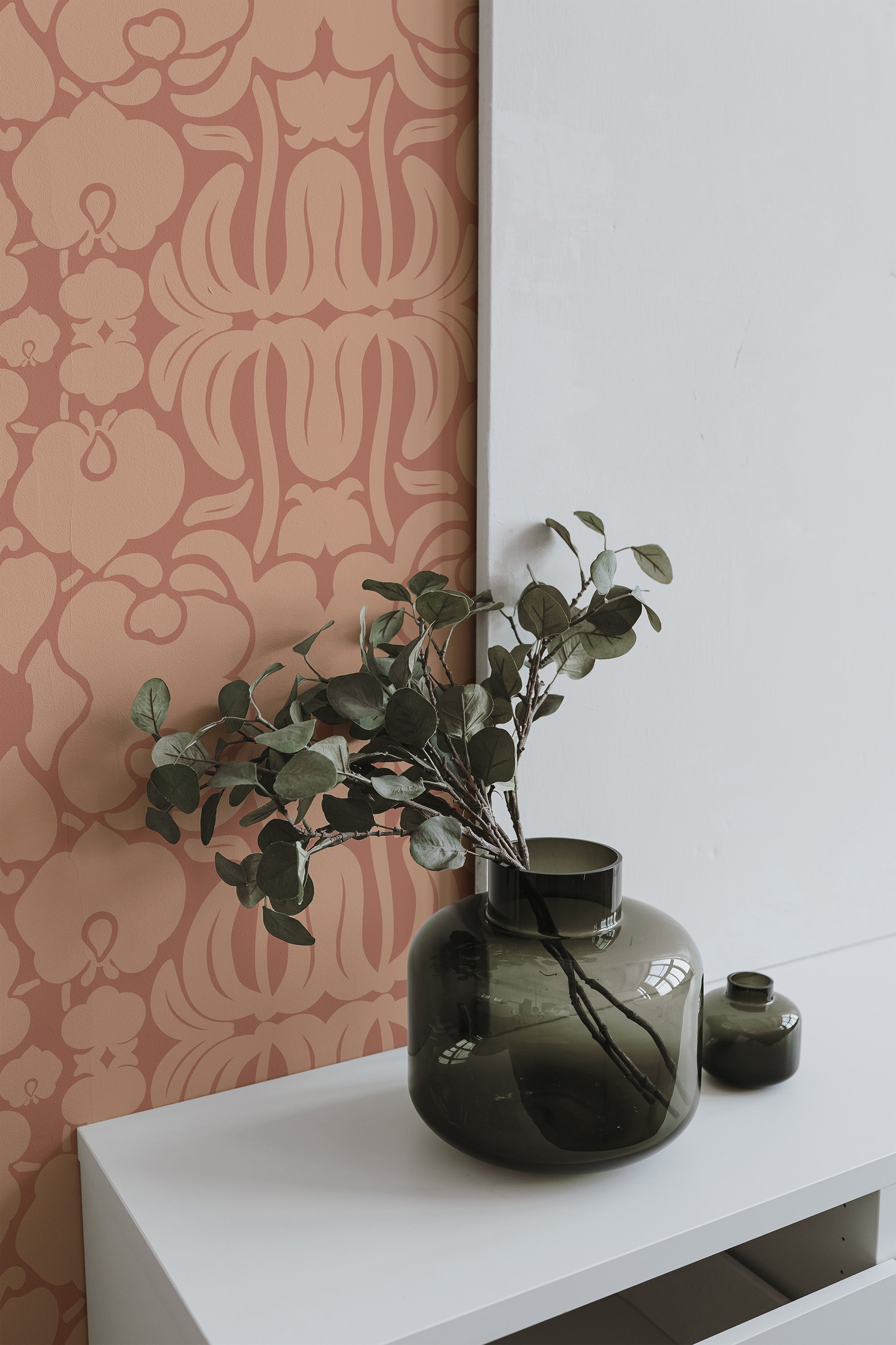Self-adhesive Victorian Peach wallpaper with ornate floral patterns on a peach-colored background, accented by a dark green vase with eucalyptus branches on a white surface.