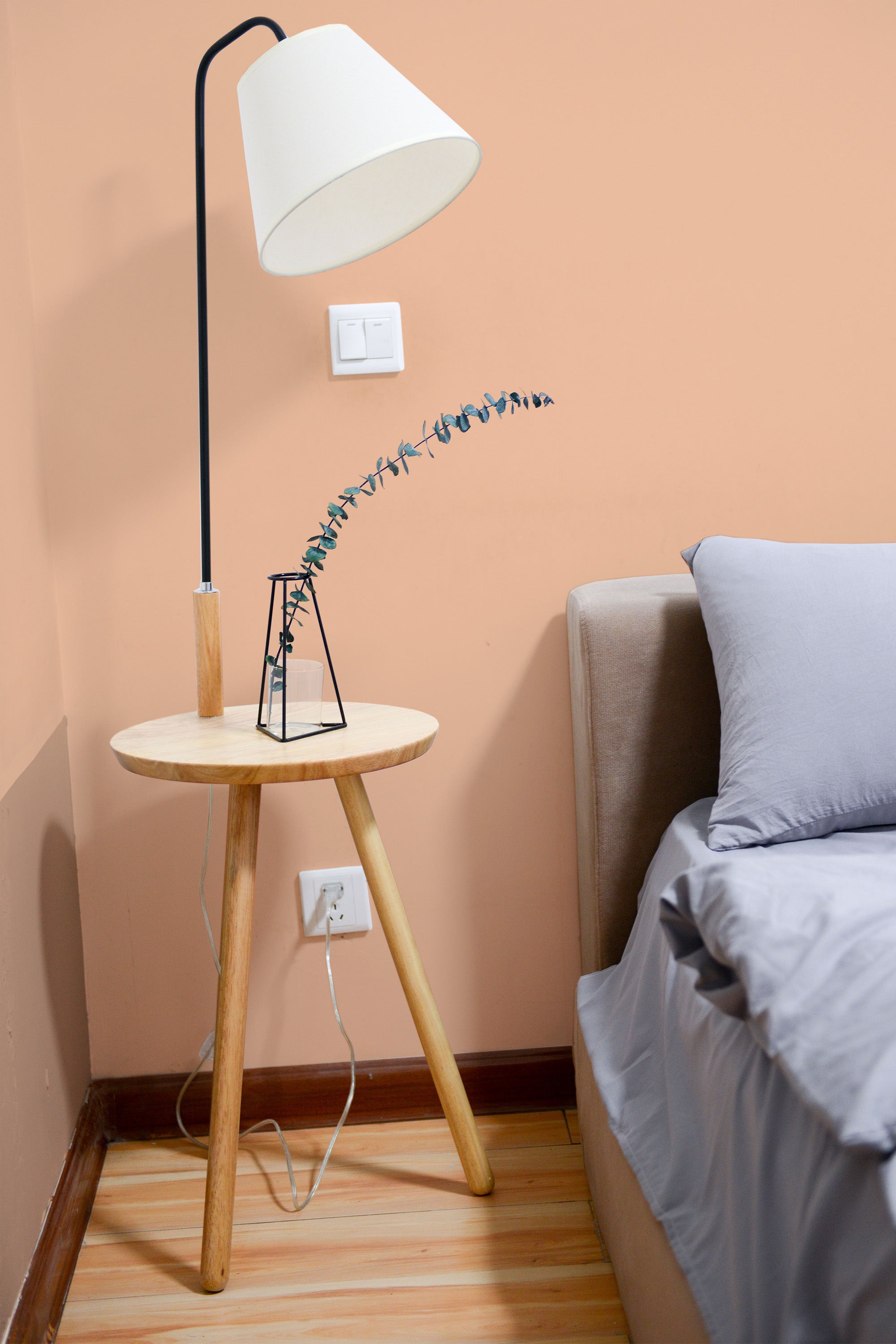 Self-adhesive solid color peach wallpaper on a room wall with a bedside table, lamp, and part of a bed visible.