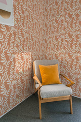 A room with a wall covered in neutral leaf bush patterned wallpaper, with a wooden chair and a yellow cushion in front, evoking a traditional wallpaper style.