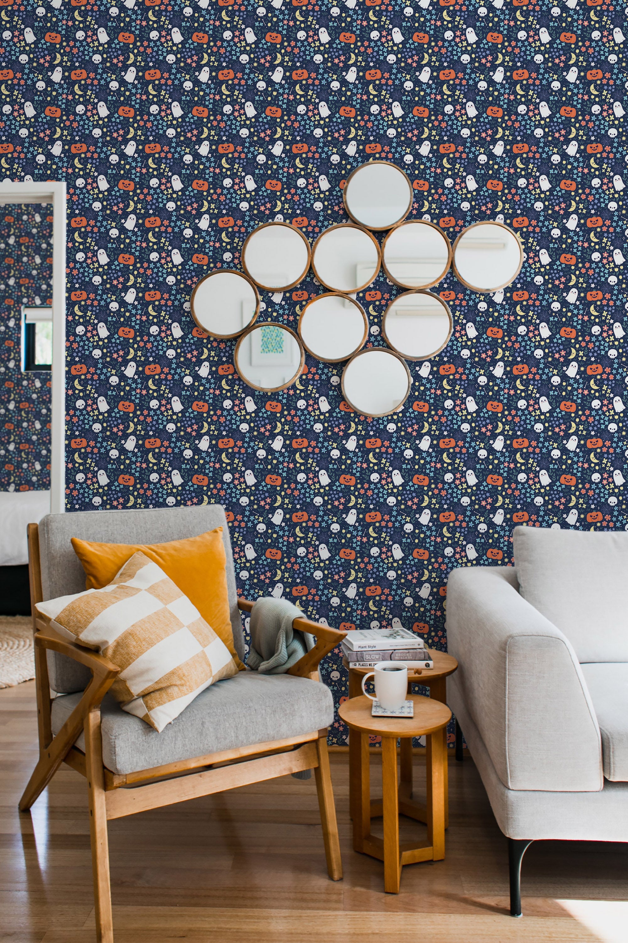 A room with navy blue Halloween-themed wallpaper with small motifs of pumpkins, ghosts, and candy corn. A wooden armchair with a yellow-striped pillow, a side table, and a white sofa are in front of the traditional wallpaper.
