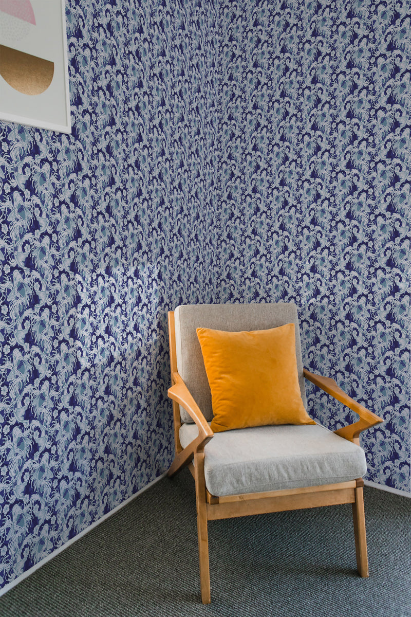 A room with blue and white nautical fish patterned wallpaper on one wall, accompanied by a wooden armchair with a grey seat and an orange pillow, embodying a traditional wallpaper style.