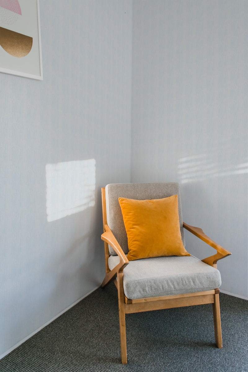 A cozy room corner with a wooden armchair and a yellow cushion against a wall covered in light blue Rainy Day themed traditional wallpaper.