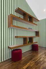 A room with green and white striped retro geometry wallpaper on one wall, accompanied by a wooden shelving unit and two red stools, reflecting a traditional wallpaper style.