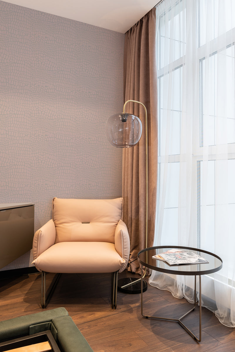 A room with Snakeskin-patterned wallpaper in muted tones, paired with a tan armchair, sheer curtains, and a glass pendant light, creating a modern twist on traditional wallpaper.