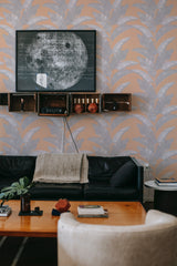 A room with walls covered in Peachy Tropical wallpaper with a pattern of large, stylized peach and green palm leaves, complementing a modern interior with a black sofa and wooden shelves, traditional wallpaper.