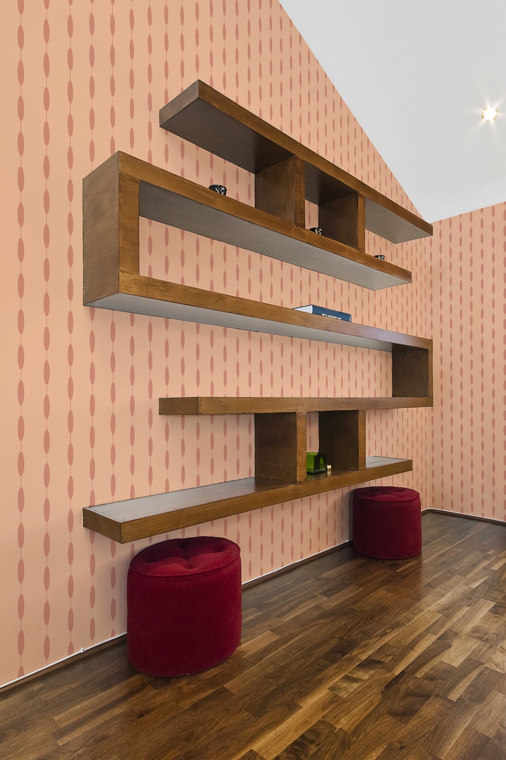 A room with Peachy Ovals wallpaper, which has a pattern of pink vertically-aligned ovals on a lighter pink background, complemented by wooden shelves and two red stools, giving a modern twist to traditional wallpaper.