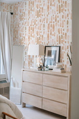 A room with walls covered in Peachy Watercolor Drops wallpaper, a pattern of irregular peach and beige droplet shapes against a light background, above a wooden dresser with a lamp and mirror, giving a soft, artistic touch to the space. Traditional wallpaper.