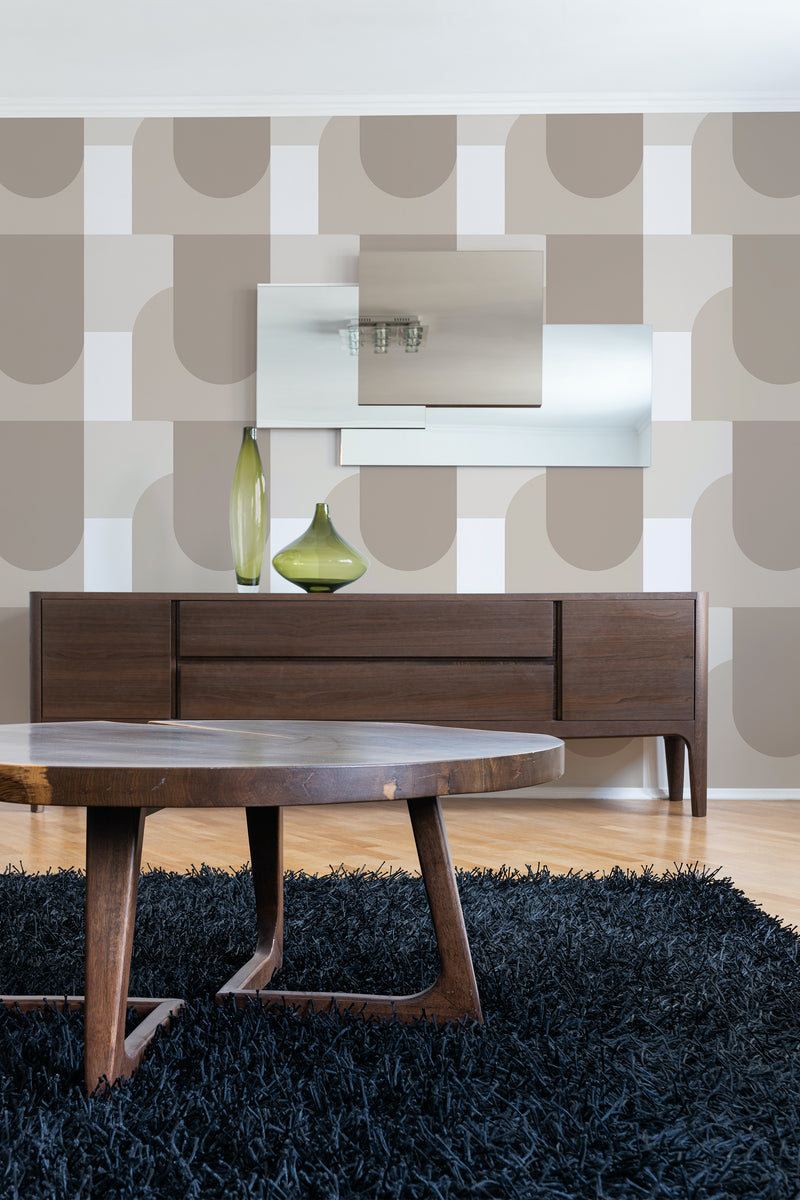 A room with neutral-toned geometric patterned wallpaper with large beige, brown, and gray overlapping circles and squares on a wall, above a wooden mid-century modern credenza and table, on traditional wallpaper.