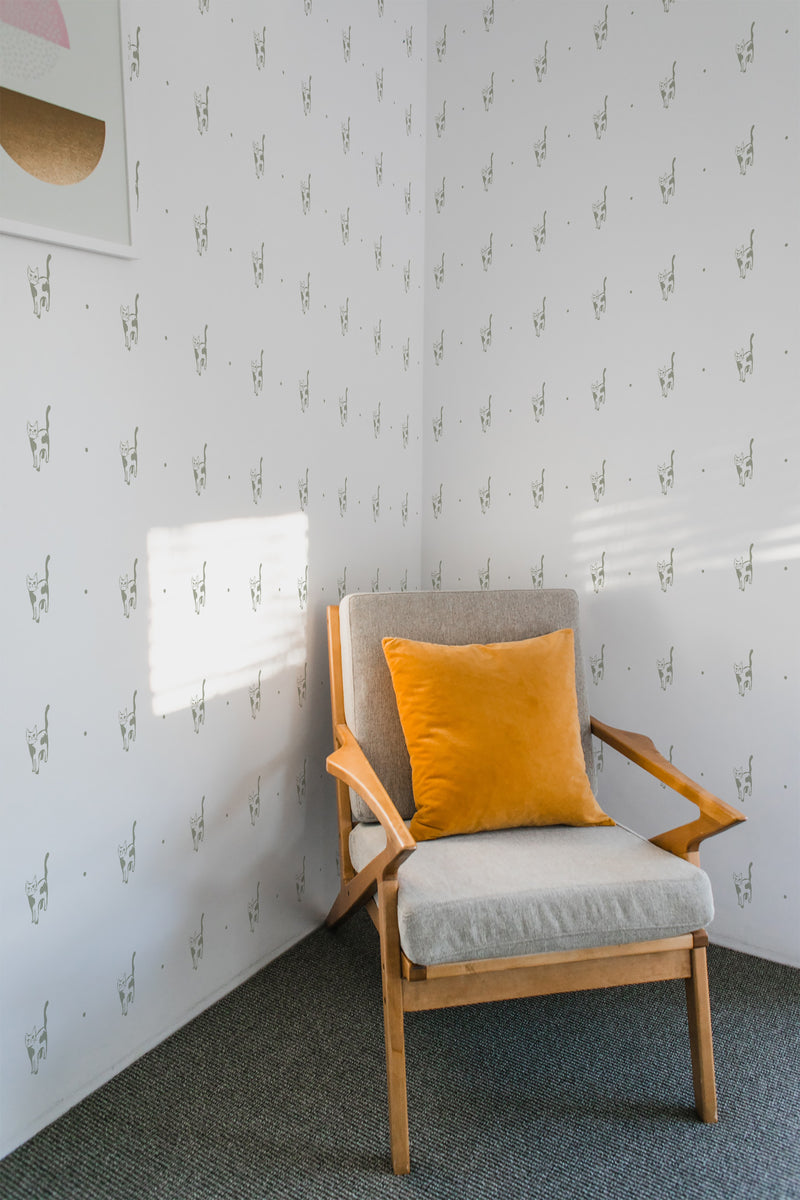 A room with white wallpaper patterned with repeated illustrations of a 'Serious Cat' motif, accompanied by a wooden armchair with a yellow cushion, reflecting a modern twist on traditional wallpaper.