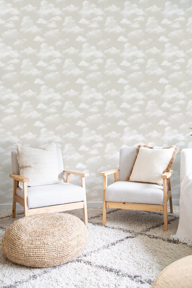 A room with light-toned Beachy Clouds in Neutral wallpaper, showing fluffy white clouds on a soft beige background, with wooden chairs and a woven ottoman, traditional wallpaper.