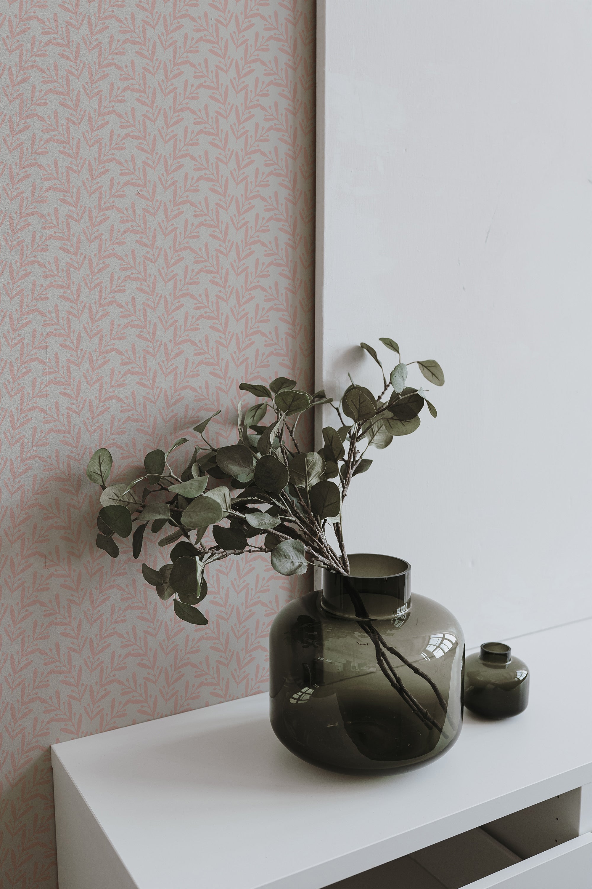 Self-adhesive wallpaper with a subtle pink background and delicate white spring leaf patterns on a wall, complemented by a dark green vase with foliage on a white shelf.