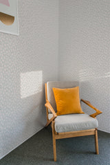 A room with bright sunlight and shadows, a wooden chair with a yellow cushion against a wall covered in small, delicate spring flower patterns on a neutral background, evoking a traditional wallpaper style.