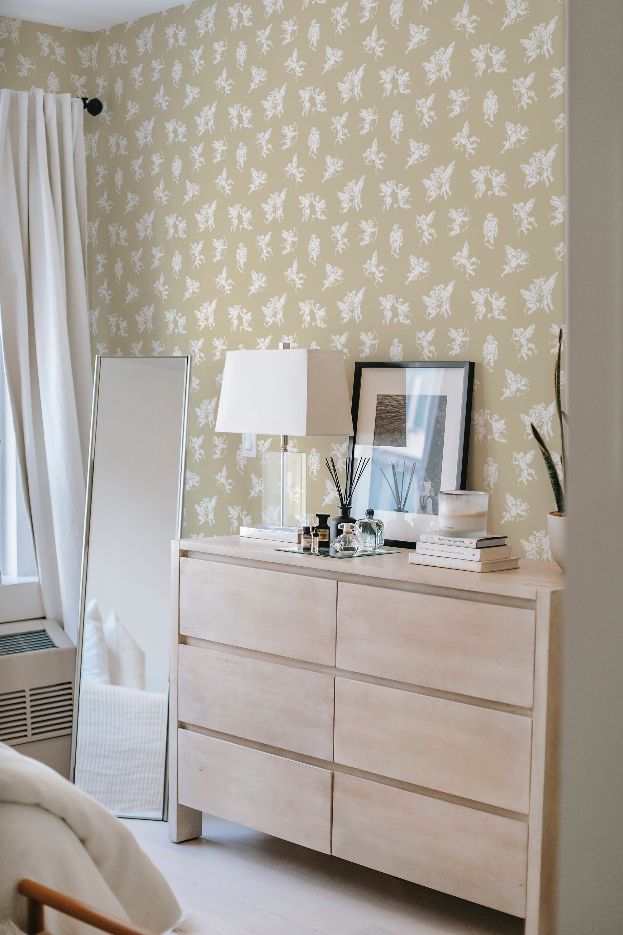 A cozy room with a dresser and lamp against a wall covered in vintage-style angel motif wallpaper in a repeating pattern, giving the space a quaint and traditional wallpaper feel.