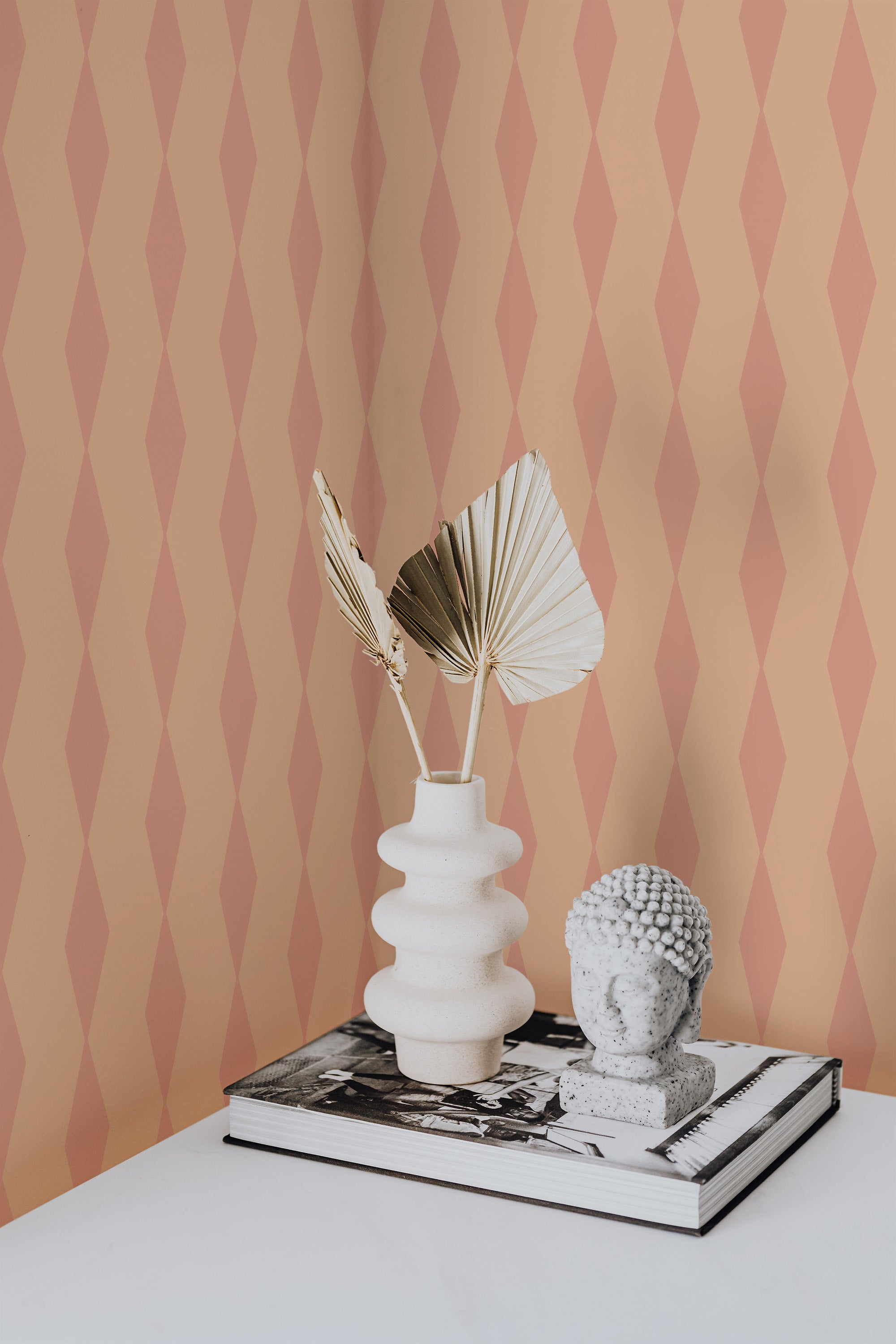 Self-adhesive wallpaper with a geometric pattern of pastel pink and beige rhombuses on a wall, complemented by a decorative vase and sculpture on a book.