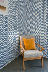 A room with blue bird patterned wallpaper on the walls behind a wooden armchair with a gray cushion and orange pillow, embodying a traditional wallpaper style.