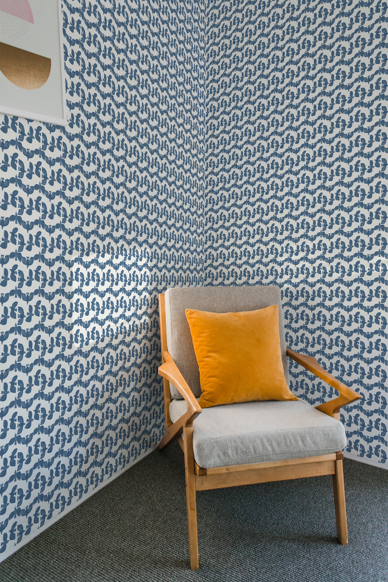 A room with blue bird patterned wallpaper on the walls behind a wooden armchair with a gray cushion and orange pillow, embodying a traditional wallpaper style.