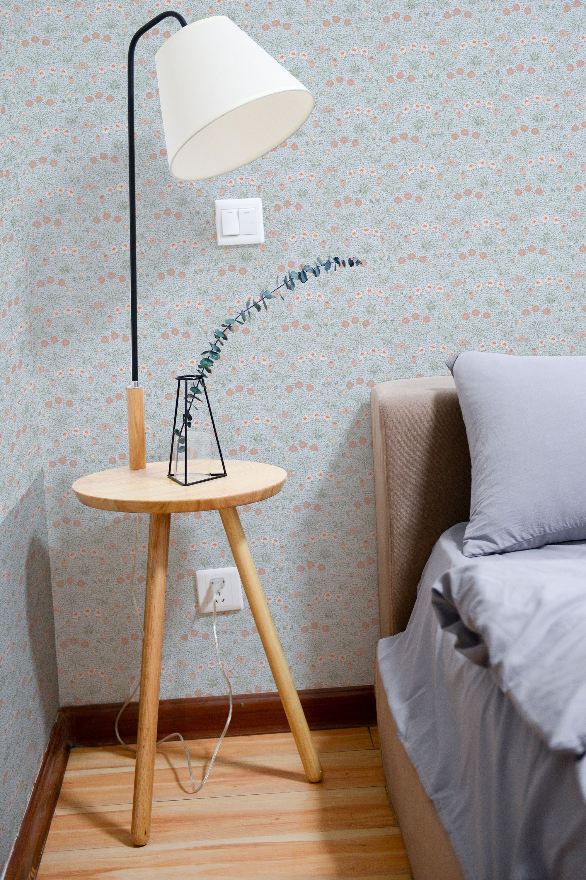Self-adhesive gray floral wallpaper on a room's wall, complemented by a simple side table, lamp, and part of a bed with gray bedding.