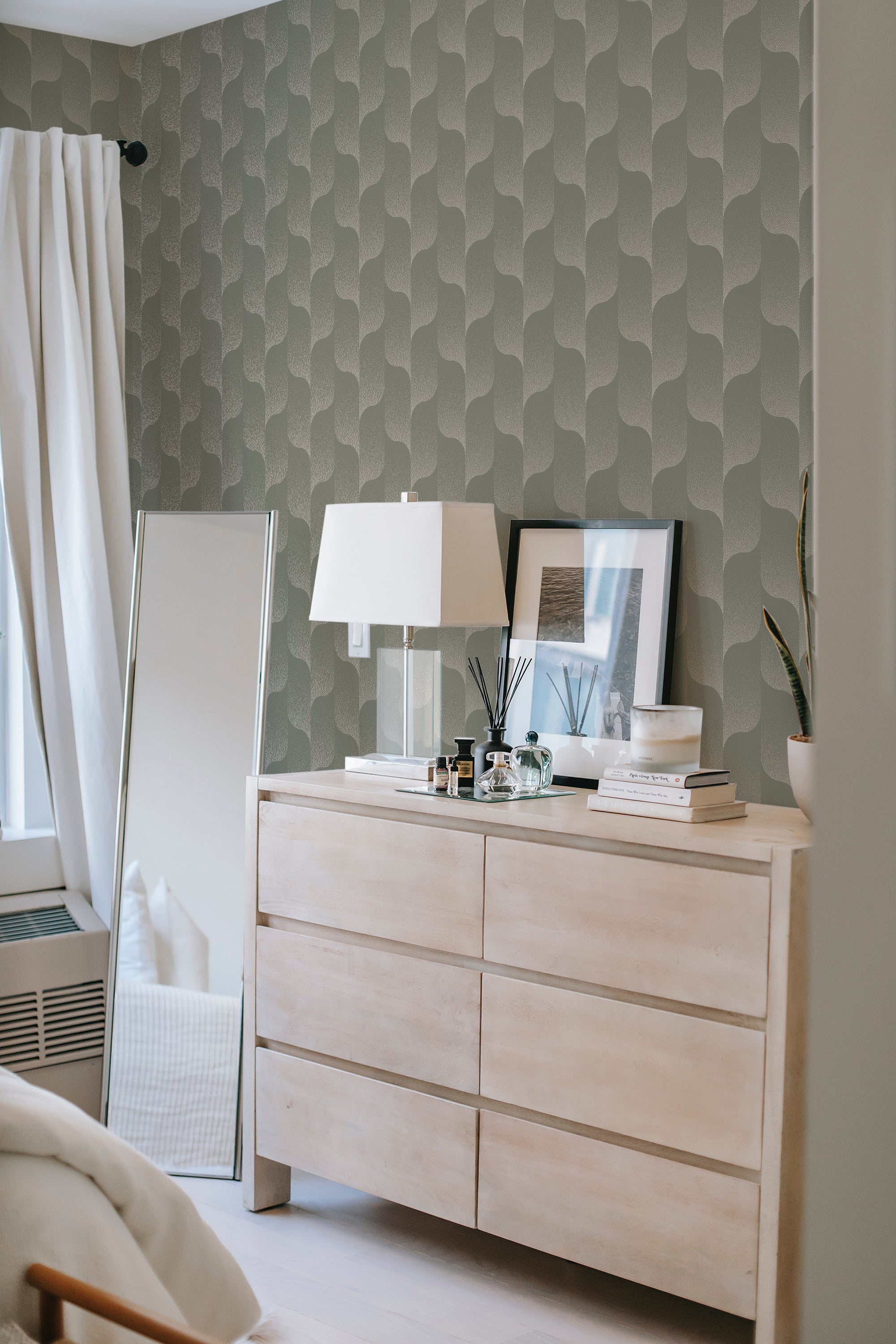 A room with a dresser and lamp in front of a wall with green-toned gradient wallpaper in a wavy design, giving a modern twist to traditional wallpaper.